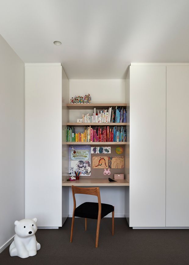 A compact, built-in study nook or children's reading and homework area set between tall white storage cabinets. Open timber shelving displays colourful books and children's artwork above a simple desk, paired with a mid-century-style wooden chair. The room has white walls and cabinetry, a dark carpeted floor, recessed ceiling lighting, and a clean, contemporary built-in design.