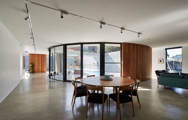 Bright open-plan contemporary interior featuring a dining area in the foreground, a curved black-framed glazed partition, polished concrete flooring, white walls and ceiling, warm timber wall panelling, and a partially visible living room with a dark teal sofa. Track lighting runs across the ceiling, and the glazing opens the room visually to an outdoor courtyard or terrace.
