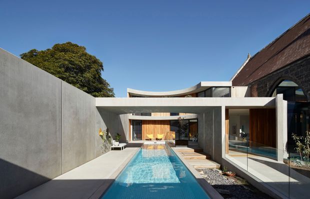 Contemporary residential courtyard viewed from outdoors, centred on a narrow lap pool enclosed by smooth concrete walls and framed by a cantilevered flat-roof structure. Glazed living spaces, warm timber joinery, pale paving, and a contrasting dark pitched roof are visible, with mature planting beyond the courtyard wall.
