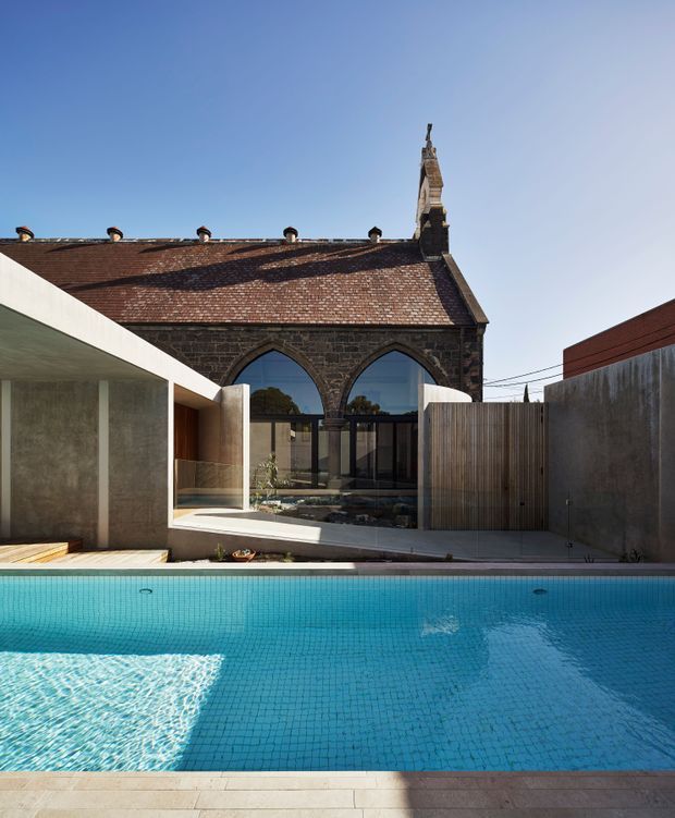 Exterior architectural photograph of a contemporary residence with a large turquoise swimming pool in the foreground. A minimalist concrete addition with broad glazing and timber detailing is set against a preserved dark stone church-like heritage building with a steep tiled roof, arched windows, and ornate stone bell-tower element. The composition highlights the contrast between historic masonry and modern extension, with paved pool surrounds and tall boundary walls.