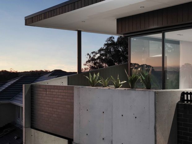 Exterior architectural view of a contemporary upper-level terrace or balcony at dusk. The composition features a flat projecting roof canopy, dark vertical timber cladding, exposed smooth concrete planter and wall surfaces, large dark-framed glazing, and integrated architectural planting. A neighbouring roof and distant tree line are visible in the background.