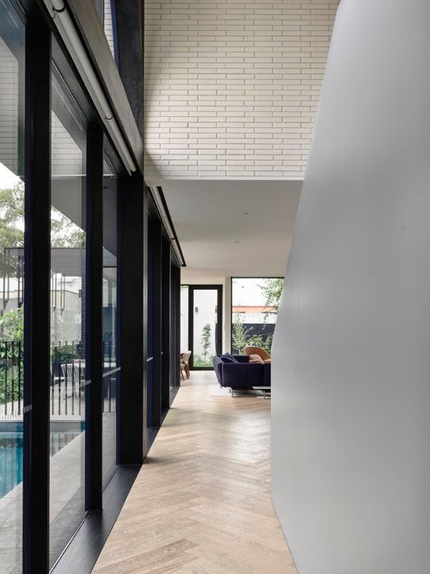 A contemporary interior corridor leading toward a living area, with full-height black-framed glazing along one side overlooking a pool and outdoor area. The hallway has pale herringbone timber flooring, smooth white walls and ceiling, and a projecting white brick-clad upper volume. A dark sofa and warm-toned lounge chair are visible at the far end.