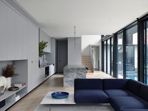 A refined open-plan contemporary interior combining a minimalist kitchen, marble island, living area, and stair hall. Full-height black-framed glazing lines the right side, bringing in abundant light and views of a swimming pool. White built-in cabinetry, pale timber flooring, a dark blue sectional sofa, and a sculptural marble island create a clean, sophisticated space.