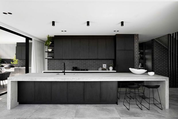 Contemporary open-plan kitchen dominated by a substantial pale stone island with integrated sink and black tap. The kitchen features full-height matte dark cabinetry, a dark brick splashback and column, black bar stools, recessed and track-style ceiling lighting, and large-format grey tiled flooring. A dining or living area is partially visible beyond the kitchen.