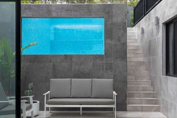 A compact contemporary courtyard or outdoor lounge with a grey upholstered bench beneath a bright blue rectangular pool or water feature set into a dark grey tiled wall. Pale tiled stairs rise alongside a grey rendered wall with black-framed glazing and wall lights. Tropical planting is visible at the edge, and the space has a refined, minimalist architectural character.