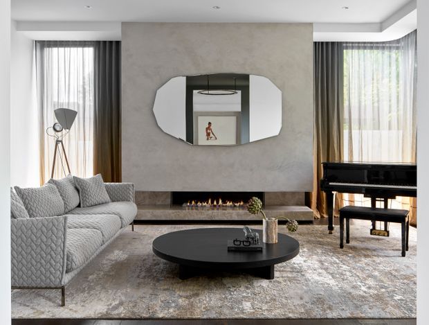 A refined contemporary living room with a large light-grey sectional sofa, black oval coffee table, textured area rug, grand piano, and a linear fireplace set into a full-height textured feature wall. Floor-to-ceiling sheer and dark drapery frame the windows, while a sculptural oval mirror and subtle recessed ceiling lights add visual interest.