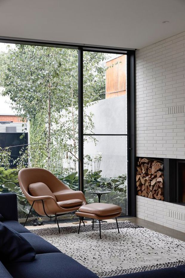 A refined living room with full-height black-framed glazing overlooking a leafy courtyard, a white painted brick feature wall, and a built-in fireplace with stacked firewood storage. Mid-century-inspired leather lounge chair and ottoman sit beside a dark sectional sofa on a large patterned rug. The space is bright, warm, and connected to greenery outside.