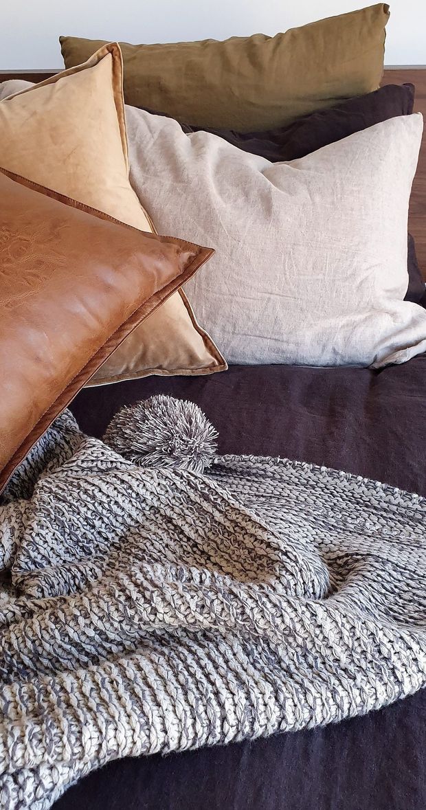 Close-up interior styling photograph of a dark upholstered sofa layered with neutral linen and leather cushions, plus a chunky grey-and-white knitted throw with a pom-pom detail. The composition emphasizes soft textiles, tactile surfaces, and a warm, relaxed lounge setting.