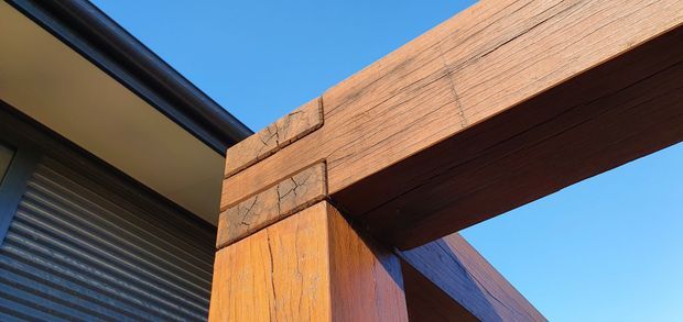Close-up exterior architectural detail showing a substantial timber post-and-beam structure with visible grain, joinery, and weathered texture beside dark metal cladding and a roller door, set against a clear blue sky.