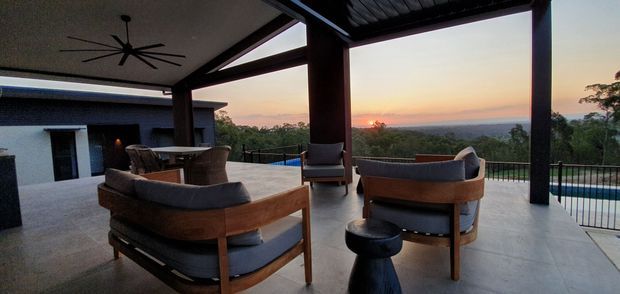 A covered outdoor lounge terrace at sunset, furnished with timber-framed upholstered lounge chairs and a small side table. The open-sided structure has dark posts, a metal roof, concrete flooring, and overlooks a landscaped setting with a swimming pool and distant water or horizon views.