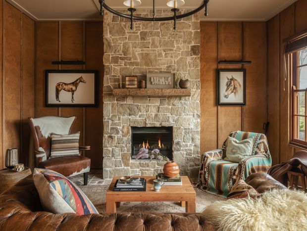 A richly furnished traditional living room centred on a rough-cut stone fireplace with an active fire and timber mantel. Dark warm wood wall panelling surrounds the fireplace, with framed equestrian artwork, leather seating, patterned upholstered armchairs, a timber coffee table, and a tufted leather sofa in the foreground. A multi-arm chandelier and a timber-framed window reinforce the cosy lodge-like character.