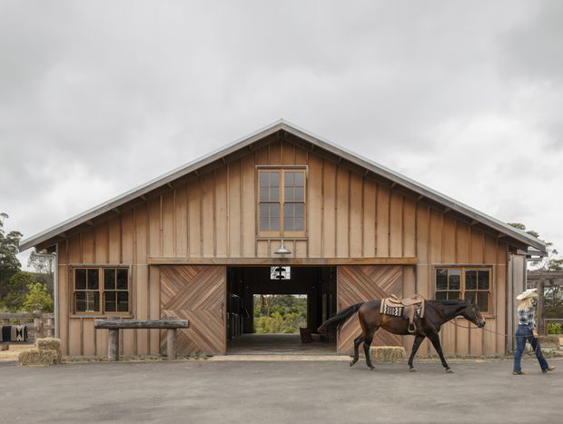 Front exterior view of a rustic timber-clad barn or stable with a symmetrical gabled roof, large central opening, cross-braced timber doors, multi-pane windows, and a horse in the foreground. The building sits in a rural-looking setting under an overcast sky.