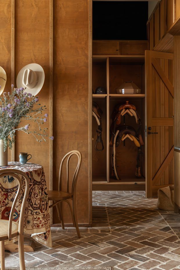 Warm, richly detailed interior showing a timber-lined entryway or mudroom with an open barn-style storage door and built-in shelving containing saddles and riding gear. A small dining table with patterned textile, bentwood chairs, hats, a vase of dried flowers, and pottery occupy the foreground. The floor is laid in a rustic herringbone brick pattern, creating a farmhouse or equestrian-inspired atmosphere.
