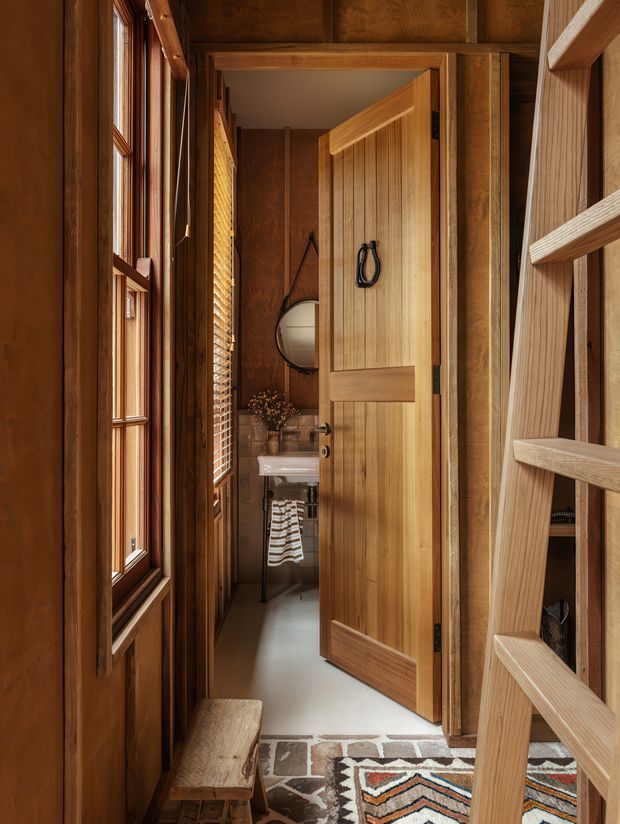 A narrow timber-lined hallway or entry passage leading to a compact bathroom. The space features warm wood wall and ceiling finishes, a multi-pane timber window with blinds, a partly open paneled timber door, and a small white wall-mounted basin visible beyond. A round mirror, towel, woven patterned rug, timber bench, and ladder-like open shelving add detail. The image has a warm, rustic cabin-like character with carefully crafted joinery.