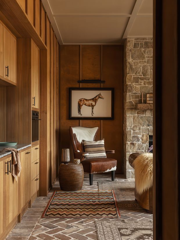 A narrow, warmly lit interior passage or sitting nook with extensive built-in timber cabinetry along the left wall, a dark timber-panelled feature wall, and a rustic exposed-stone wall on the right. A leather armchair with a light hide draped over its back sits beneath framed horse artwork, accompanied by a woven side table and layered patterned rugs. The floor is finished in irregular stone or brick-like paving, creating a rich lodge-inspired atmosphere.