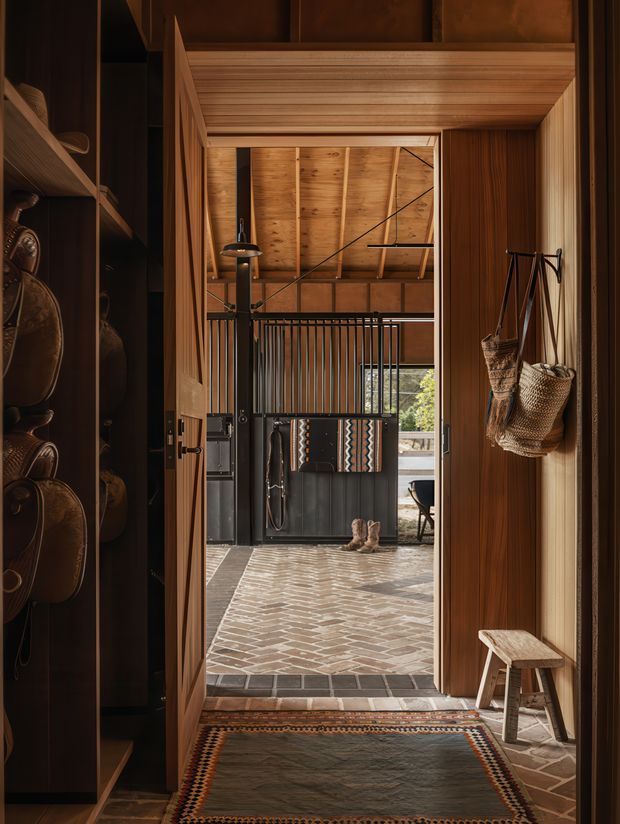 A warm, richly timber-lined entryway opening into a covered outdoor or semi-enclosed foyer. The view features a heavy timber-framed doorway, dark built-in shelving with leather goods, woven hanging bags, a small timber stool, patterned runner, brick-paved floor, black metal balustrade, and a timber-clad pitched roof beyond. The atmosphere is rustic, earthy, intimate, and highly crafted.