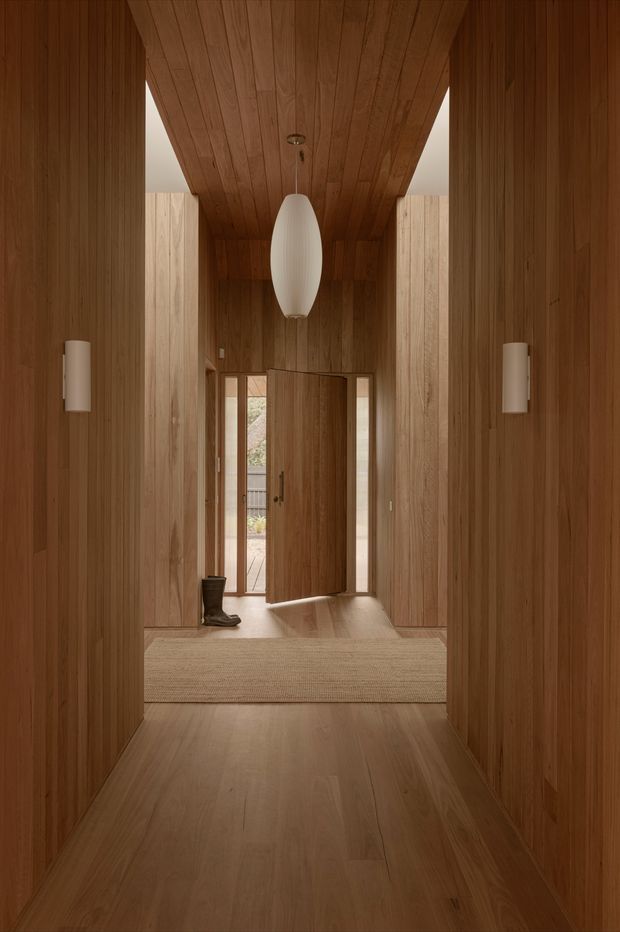 A warm, timber-lined residential entry hall viewed from inside. The walls, ceiling, built-in door, and floor are clad in rich natural wood, creating a continuous enveloping interior. A large sculptural white pendant hangs centrally, with matching slim wall sconces on both sides. An open timber front door leads to a small exterior deck, while a textured neutral runner and a pair of boots sit near the threshold.