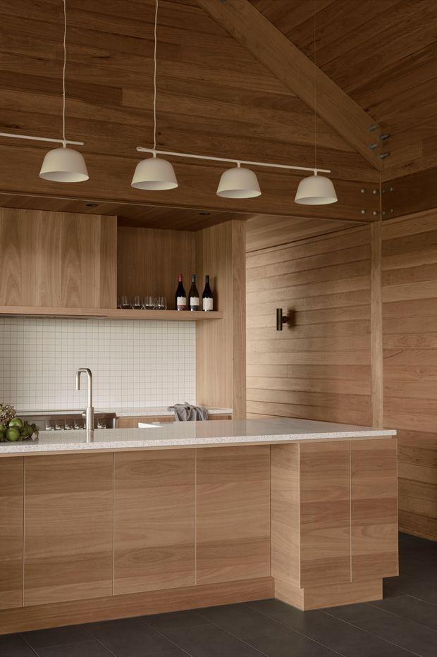 Warm, wood-lined kitchen with extensive built-in timber cabinetry, a white square-tile splashback, pale speckled stone island benchtop, dark tiled flooring, and four suspended cream pendant lights. The room includes a sink, kitchen work area, open shelving with wine bottles and glassware, and a strongly expressed timber ceiling and wall structure.