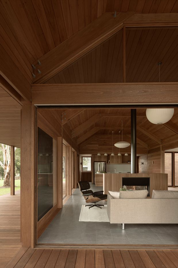 A warm, timber-lined open-plan interior viewed through large glazed openings, featuring a vaulted exposed-beam ceiling, wood wall panelling, grey tiled flooring, a contemporary lounge with a cream sofa and leather lounge chair, and a built-in fireplace partition separating the living area from a kitchen with an island and pendant lights. A timber deck and garden are visible outside.