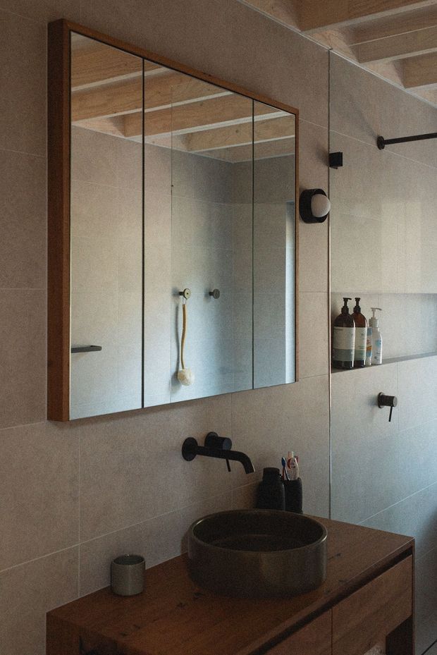 A warm, contemporary bathroom vanity with a round dark vessel basin, wall-mounted matte-black tapware, and timber cabinetry. Large mirrored cabinets with dark timber framing occupy the wall above the vanity, reflecting the tiled shower area and a timber-lined ceiling. Neutral large-format wall tiles, integrated shelving, toiletries, and a black wall light complete the understated, natural-toned scheme.