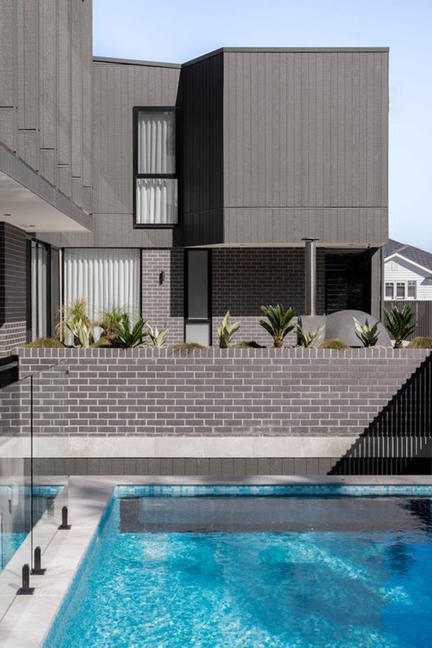 A contemporary multi-level residential facade with grey vertically textured cladding, dark brickwork, black-framed glazing, and a recessed entrance. A raised landscaped terrace sits above a swimming pool, with a stair and dark metal balustrade descending beside the pool. The composition highlights the building exterior, pool area, retaining walls, and strong geometric forms.