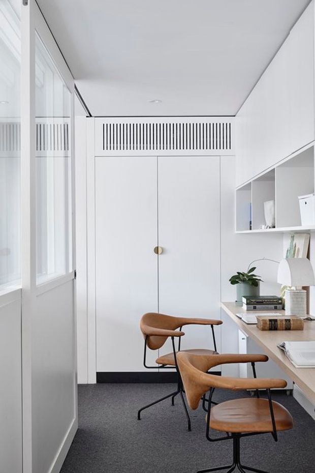 A bright, compact home office with a minimalist white built-in storage wall, integrated desk, open shelving, and concealed cabinetry. Two sculptural tan leather-and-black metal chairs sit at the desk. Full-height glazing and a glazed door bring in abundant natural light, while dark carpet provides contrast against the predominantly white interior.