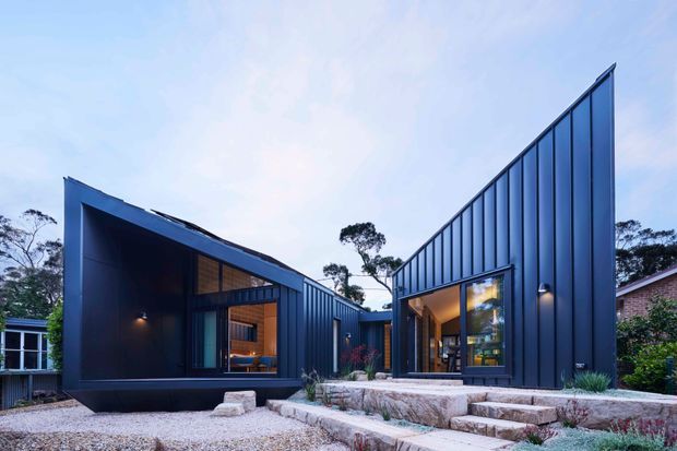 Exterior architectural photograph of a contemporary dark-clad residence composed of sharply angled, asymmetrical roof forms. The home features deep blue-black vertical metal cladding, extensive glazing, warm interior lighting, stepped stone terraces, gravel landscaping, and established trees surrounding the site.