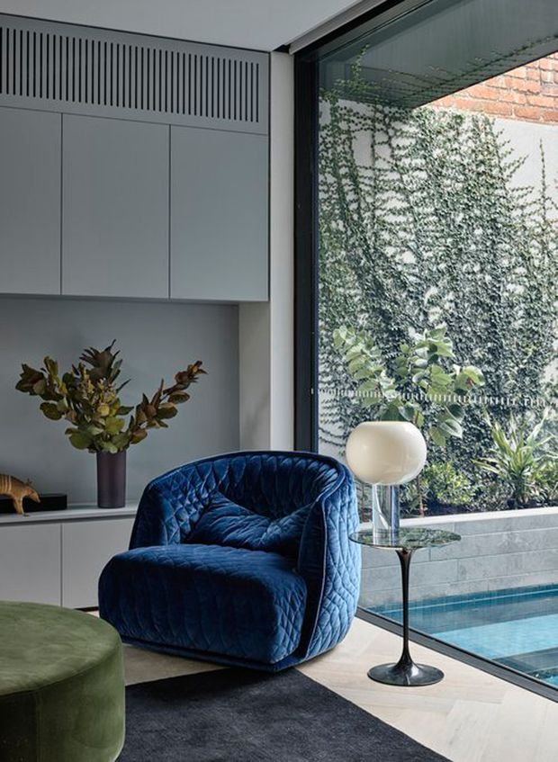 A refined modern interior corner with a deep blue quilted lounge chair beside a small glass-topped side table and spherical lamp. White built-in cabinetry with integrated vertical ventilation detailing lines the wall, while full-height glazing overlooks a planted courtyard and narrow pool. A round olive-green ottoman and dark rug add contrast to the pale timber flooring.