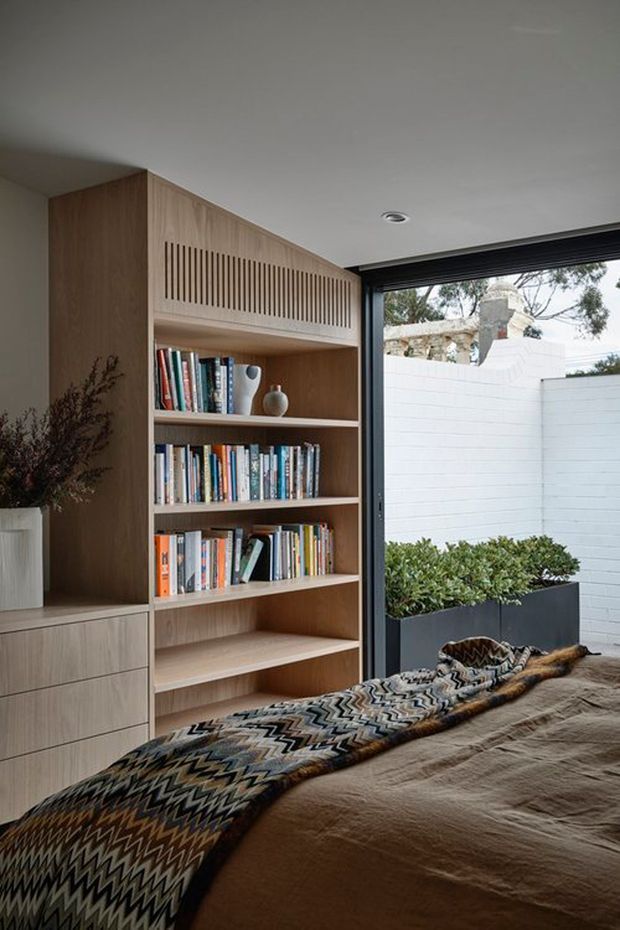 A warm, contemporary bedroom corner featuring a full-height built-in timber bookcase and integrated drawers. The open shelving contains books and small decorative objects, with a slatted timber section above. A bed with a textured patterned throw occupies the foreground, while large dark-framed glazing opens toward a small planted courtyard and white masonry boundary wall.