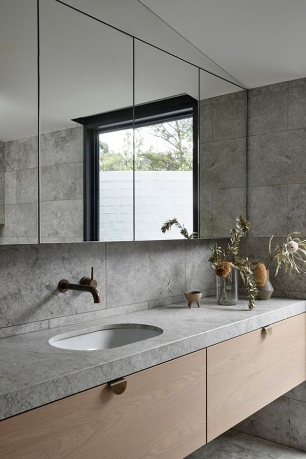 Contemporary bathroom vanity featuring a floating light timber cabinet with rounded bronze pulls, an integrated oval basin, grey stone benchtop and matching large-format grey wall tiles. A wall-mounted bronze tap sits beneath a wide mirrored cabinet, with a dark-framed window and restrained dried botanical styling.