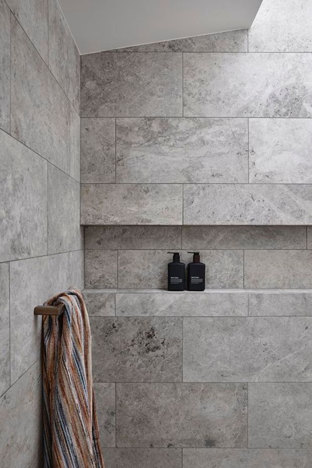 Close-up of a minimalist shower wall clad entirely in large-format grey stone-look tiles, with a recessed shelf holding two dark bottles. A timber towel rail with a striped towel is visible at the left. The space has a clean, restrained contemporary feel with subtle natural stone variation.