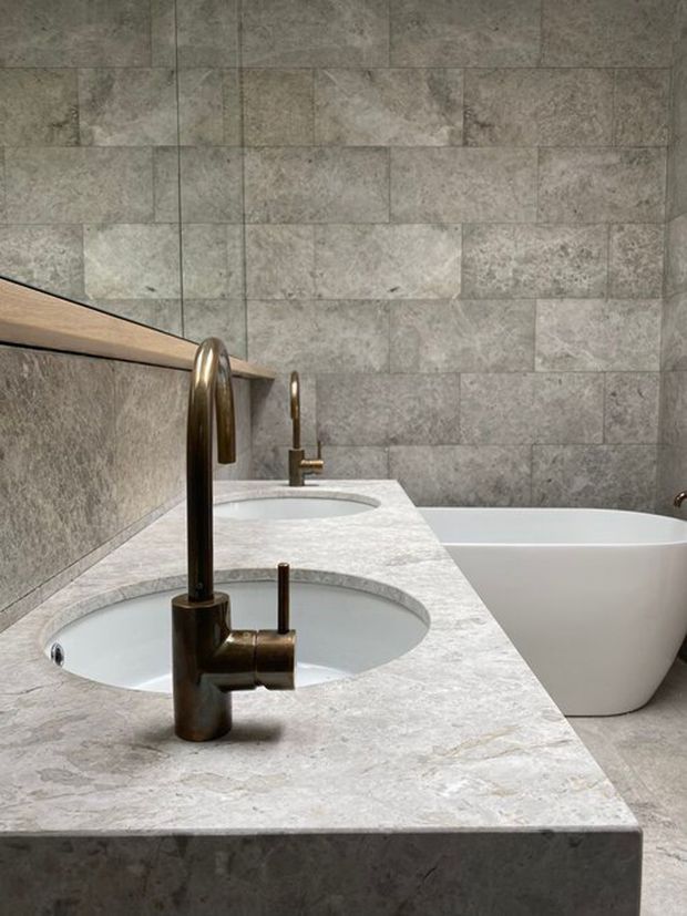 Close-up view of a refined bathroom featuring a long pale stone vanity with two integrated oval basins, dark bronze curved faucets, a freestanding white bathtub, and large-format grey stone-look wall tiles. The space has a restrained, luxurious material palette and clean contemporary detailing.