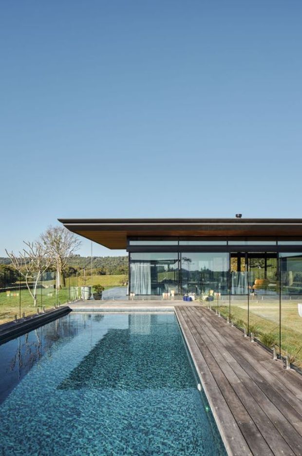 A modern single-storey residence viewed from beside a rectangular swimming pool. The image features a broad flat roof with deep timber-toned eaves, extensive floor-to-ceiling glazed walls, and a timber poolside deck. The landscaped rural setting includes lawn and trees, while the pool and reflective glazing dominate the foreground and house facade.
