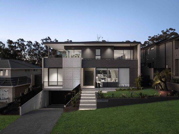 Contemporary multi-level urban residence viewed from the street at dusk. The facade combines dark brickwork, smooth light concrete panels, extensive glazing, recessed balconies, and a broad flat roof. A stepped front entry, lower-level garage access, landscaped garden beds, lawn, and neighbouring homes are visible.