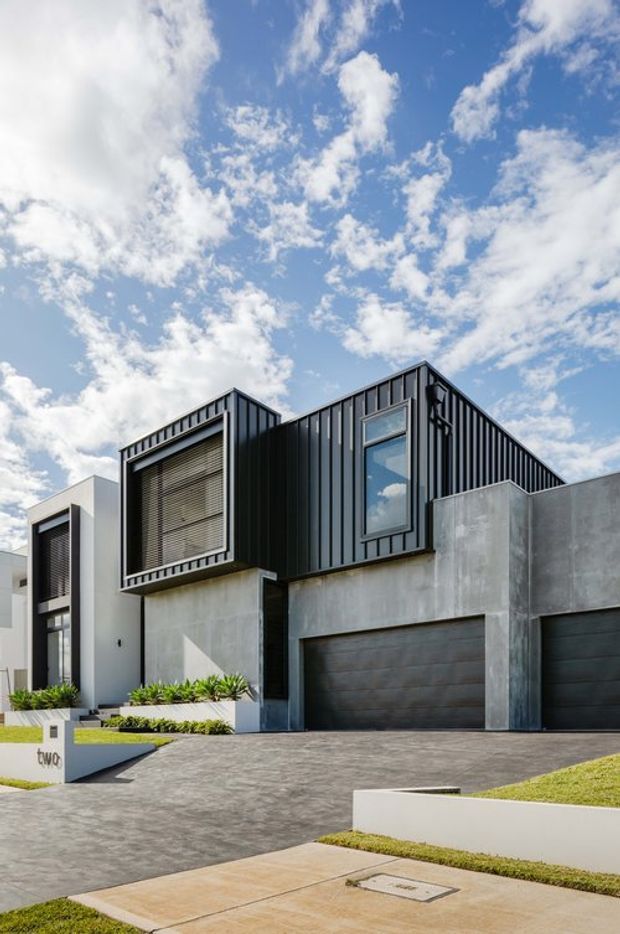 Modern multi-level residential townhouse facade viewed from the street, featuring dark vertically ribbed metal cladding on the upper storey, exposed grey concrete garage surrounds, large dark garage doors, and contrasting white rendered sections. The frontage includes stepped landscaped planters, a paved driveway, and tall rectangular windows under a bright blue sky.