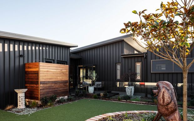 Exterior view of a contemporary dark-clad residential building arranged around a landscaped courtyard. The facade features vertical charcoal cladding, pitched roof forms, narrow windows, a timber privacy screen, artificial turf, raised garden beds, and a sculptural tree trunk or feature element.