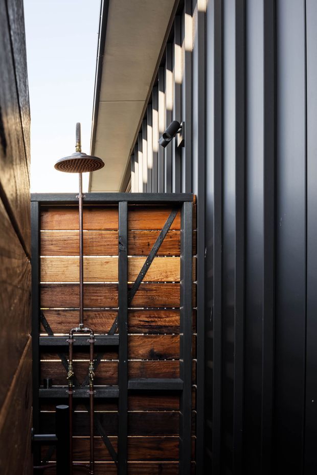 A narrow contemporary outdoor shower enclosure between dark walls. The shower features exposed copper plumbing and a round rainfall showerhead, backed by a warm reclaimed timber screen framed with black metal bracing. A dark vertical metal-clad wall and simple black exterior light create a strong industrial character.