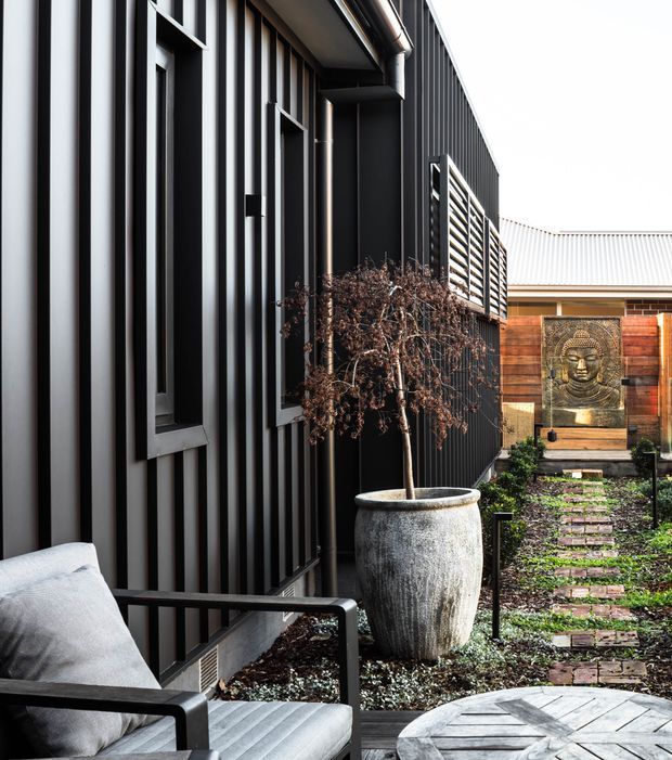 A narrow landscaped courtyard beside a contemporary dark-clad building, featuring vertical ribbed metal wall cladding, a black-framed window, an outdoor armchair, a large weathered planter, brick stepping stones, raised garden beds, and a timber privacy wall with a Buddha relief feature.