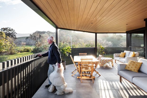 A covered, open-sided outdoor terrace with a warm timber-lined ceiling, dark vertical balustrades, large glazed openings, and light tiled flooring. The space is furnished with a timber dining table and chairs, lounge seating, striped outdoor chairs, and yellow accent cushions, overlooking a landscaped, bushy setting.