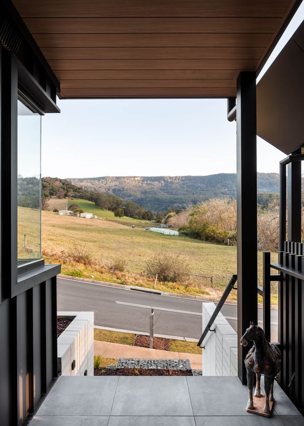 A covered entry porch or balcony looks out over a rural valley landscape. The space features a warm timber-lined ceiling, dark structural columns and window frames, large glass panels, grey tiled flooring, and a short stair descending to the road. A sculptural horse is positioned at the edge of the entry, while the surrounding landscape shows rolling farmland and wooded hills.