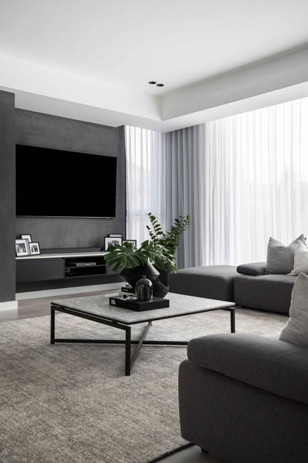 A refined contemporary living room in a restrained monochromatic palette. The space features a large grey sectional sofa, slim black metal coffee table with a pale stone-like top, textured grey carpet, a wall-mounted television set into a dark grey feature wall with low built-in media cabinetry, and full-height sheer curtains providing soft natural light. Indoor plants and framed photographs add subtle warmth.