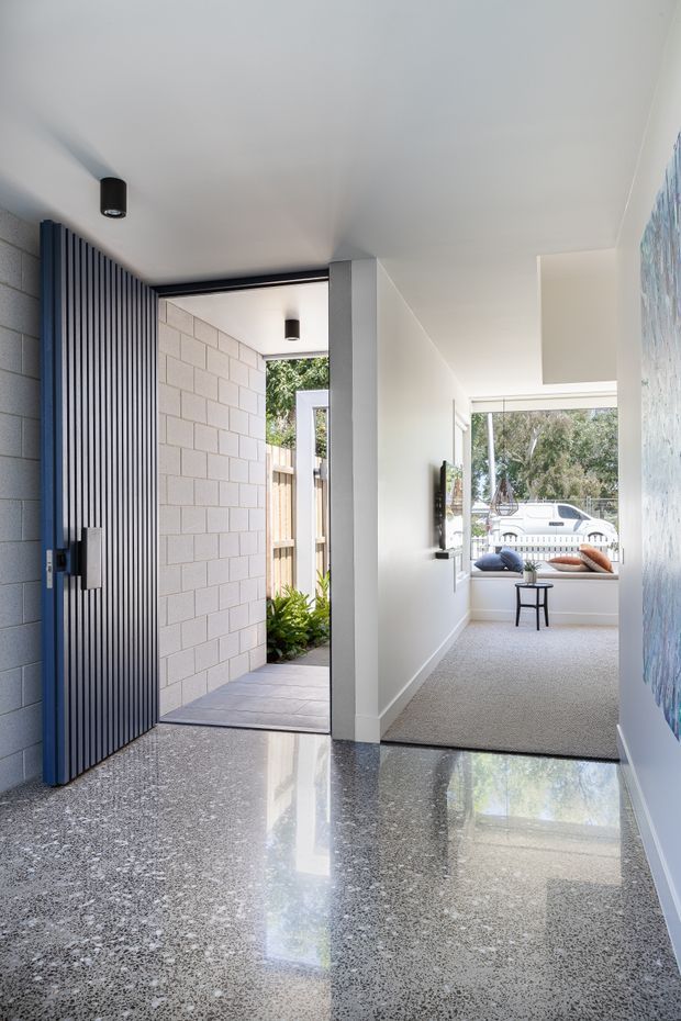 A bright contemporary residential entry hall with a large dark timber slatted front door, polished terrazzo flooring, white walls, and a view through to a carpeted sitting area and outdoor garden. A white masonry feature wall, black cylindrical pendant lights, recessed niches, and large windows add architectural interest.