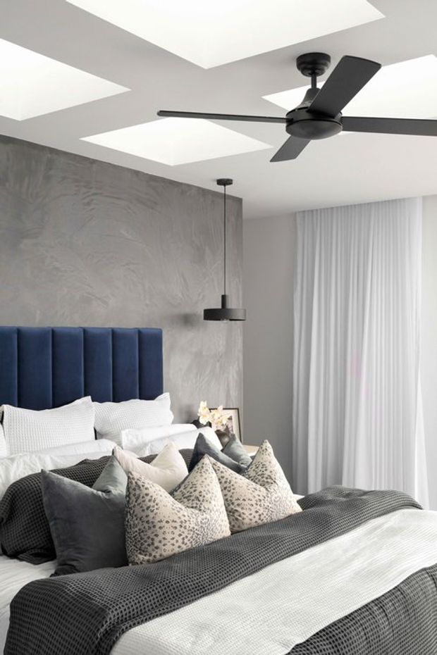 A contemporary bedroom with a large bed dressed in layered charcoal, white, and patterned cushions and bedding. The bed has a tall dark navy upholstered panel headboard against a textured grey feature wall. A black pendant light hangs beside the bed, while a large black ceiling fan and geometric recessed ceiling coffers dominate the upper portion of the room. Full-height white sheer curtains cover the window, creating a bright, refined interior.
