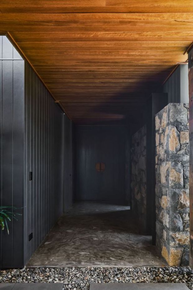 A dark, narrow covered entry passage with vertical black metal wall cladding and doors on the left, a rough natural-stone wall on the right, and a warm timber-lined ceiling overhead. The concrete-look floor transitions to a strip of rounded river stones at the threshold, creating a dramatic, tactile entrance sequence.