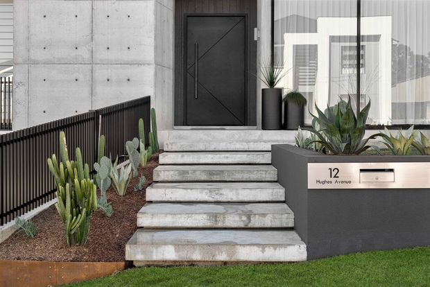 Modern residential front entry with a broad concrete staircase leading to a dark geometric entry door. The facade combines exposed concrete panels, dark metal railings, large glazed frontage, and a dark built-in planter/mailbox feature. Drought-tolerant architectural planting lines the steps and entry, with a small lawn in the foreground.