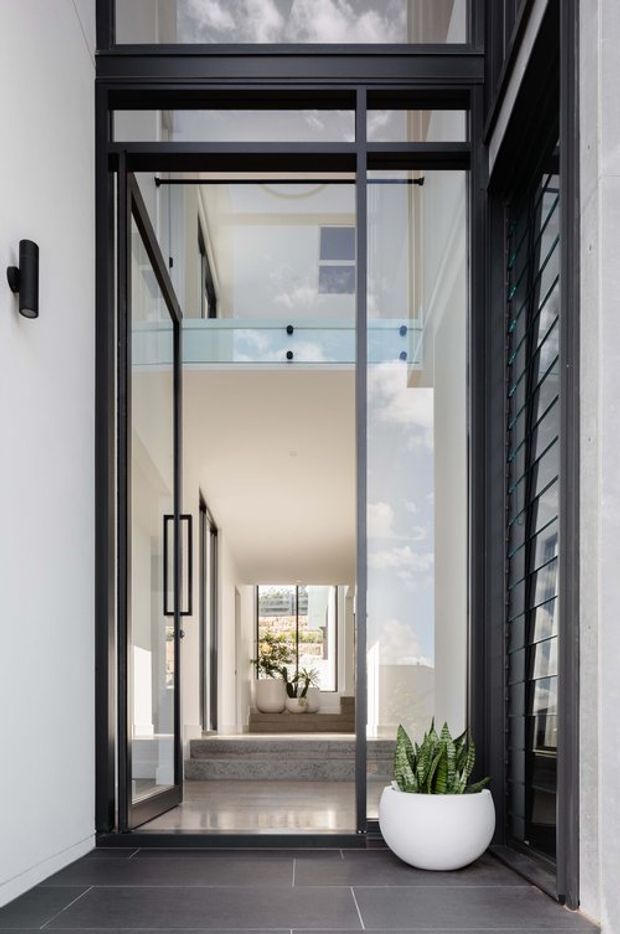 A contemporary residential entryway viewed from outside, featuring a tall black-framed glazed door and transom opening into a bright double-height interior. The entry has dark large-format floor tiles, white walls, slender black metal framing, a glass balustrade, floating or stepped internal landing, and potted plants. Strong vertical glazing and reflected sky create an airy, refined architectural feel.