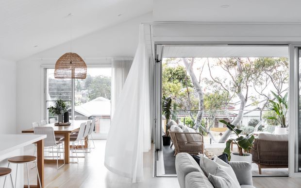 Bright open-plan interior combining a white kitchen, dining area and living space, opening onto a glazed balcony or terrace. The room features pale timber flooring, white cabinetry and walls, a woven rattan pendant light, contemporary dining furniture, indoor plants and abundant natural light through large sliding glass doors.