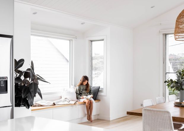 Bright, airy open-plan kitchen and dining area with white cabinetry, a built-in window seat, large white-framed windows, pale timber flooring, and a timber dining table. A child is seated on the window bench, while plants and a woven pendant add natural texture.