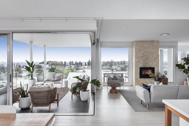 Bright contemporary open-plan living area with floor-to-ceiling glazing, a balcony view, pale timber flooring, white upholstered seating, indoor plants, and a stone-clad fireplace. The space includes a lounge area and an adjacent sunlit seating or dining zone separated by large sliding glass doors.