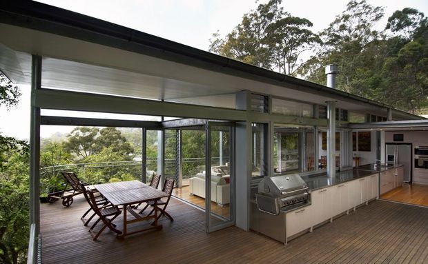 A contemporary open-plan outdoor entertaining area connected to an indoor kitchen, with a covered timber deck, dining table and chairs, lounge seating, built-in barbecue, stainless-steel kitchen appliances, extensive glazing, and a leafy wooded outlook. The low-pitched roof, slim dark framing, and indoor-outdoor layout create a relaxed modern setting.